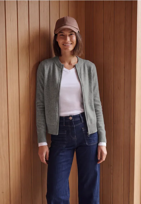 Frau vor Holzpaneelen, trägt Jeans, weißes Shirt, graue Jacke, Mütze, lächelt.