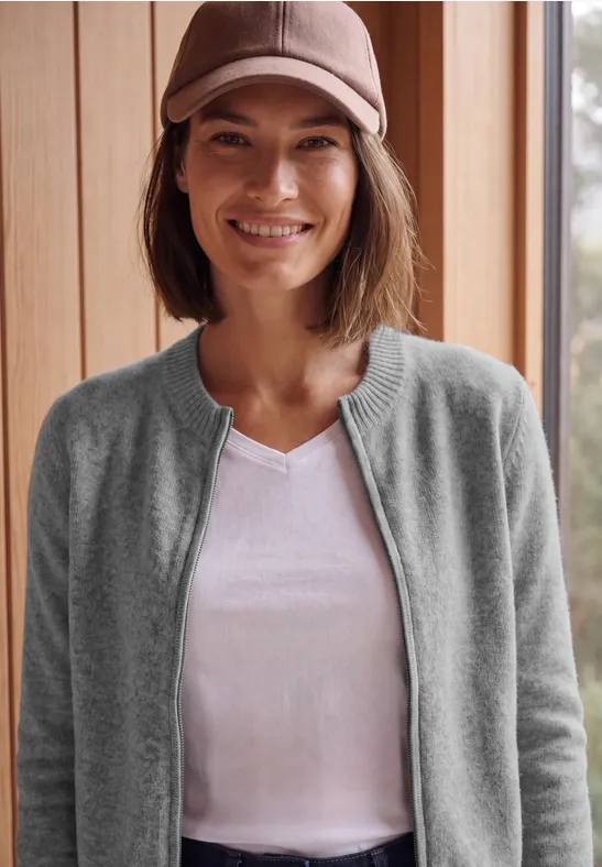 Eine lächelnde Frau mit Basecap, weißem T-Shirt und grauem Cardigan vor Holzwand.