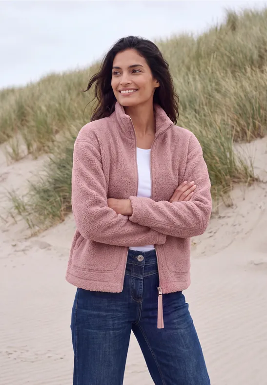Une femme souriante en veste polaire rose et jeans, bras croisés, se tient sur la plage devant des dunes.