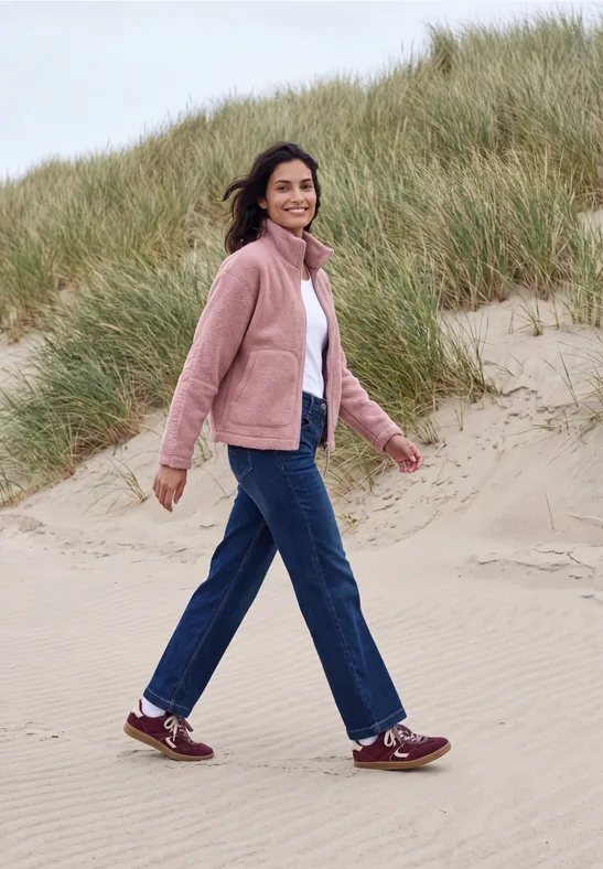 Femme en veste rose marchant sur des dunes de sable, arborant un sourire.