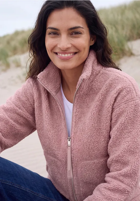 Femme souriante en polaire rose et jean sur la plage.