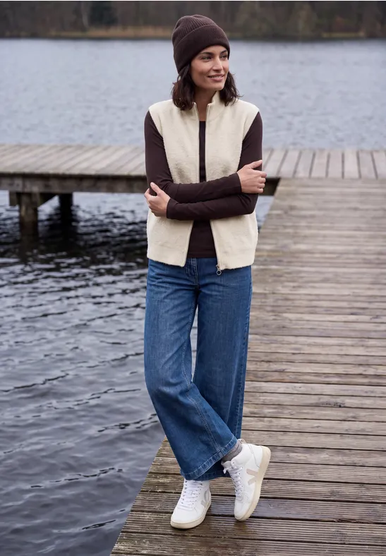 Frau in Beanie, Beigefleeceweste, braunem Pullover, Jeans und weißen Sneakern steht auf einem Steg am See.