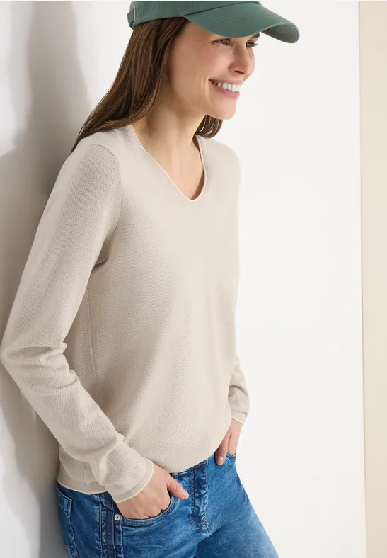 Frau lehnt lässig an einer Wand, trägt helles V-Ausschnitt-Shirt, Jeans und Baseballcap.