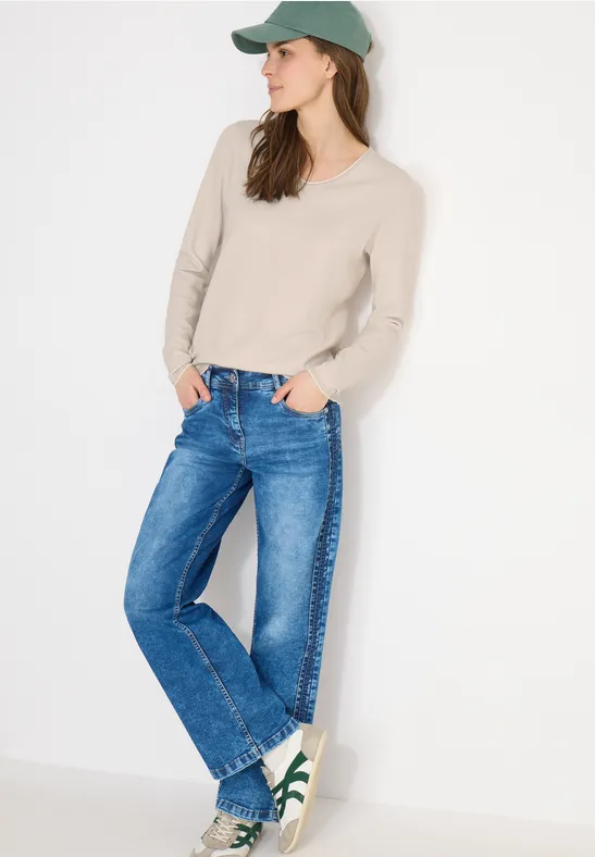 Frau in Mütze, hellem Pullover, blauen Jeans und Sneakern vor weißem Hintergrund