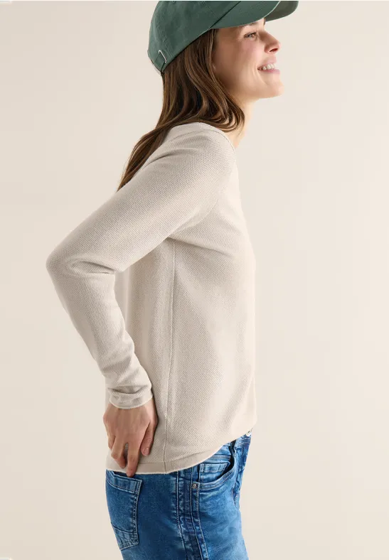 Frau in Seitenansicht trägt weißen Pullover, blaue Jeans und grünes Cap, Hände hinter dem Rücken.