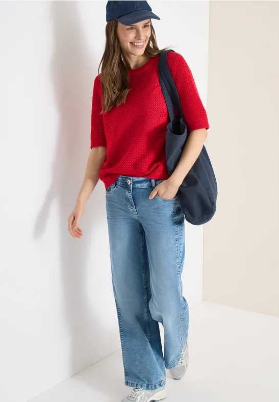 Frau in rotem Oberteil, blauen Jeans und schwarzer Tasche vor weißem Hintergrund.