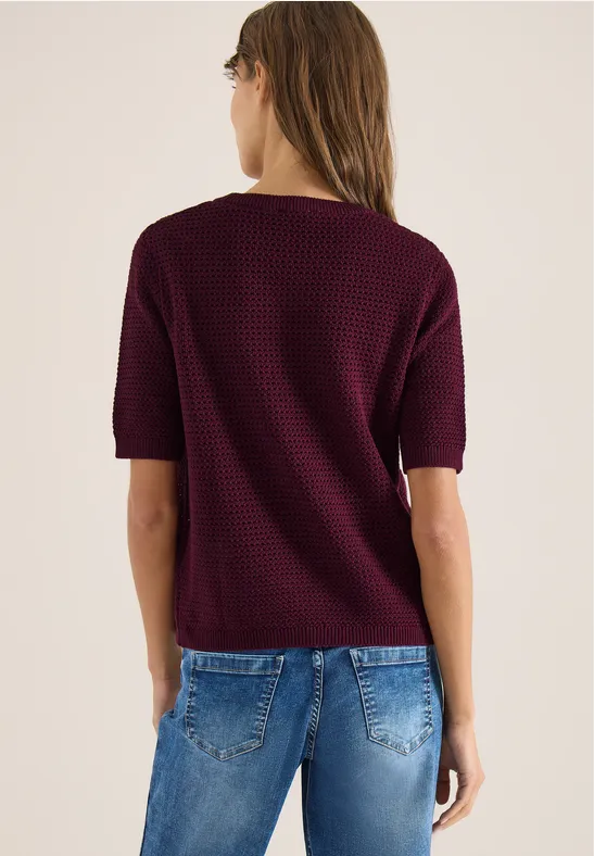 Frau von hinten in bordeauxfarbenem Kurzarm-Pullover und blauen Jeans vor hellem Hintergrund.