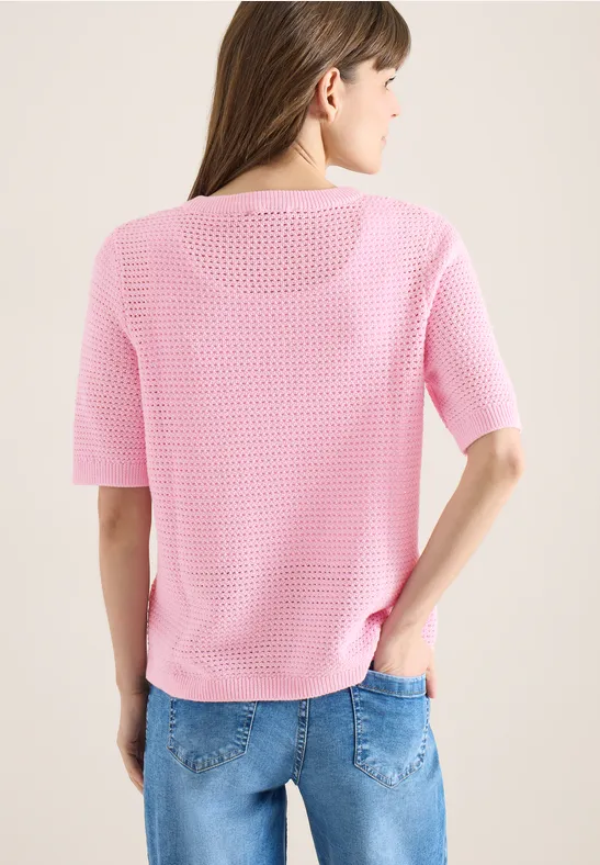 Rückenansicht einer Frau in rosa Strickshirt und blauen Jeans vor hellem Hintergrund.