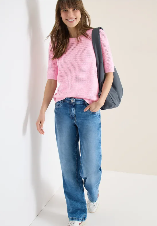 Frau in pinkem Shirt und blauen Jeans mit Schultertasche, stehend vor hellem Hintergrund.