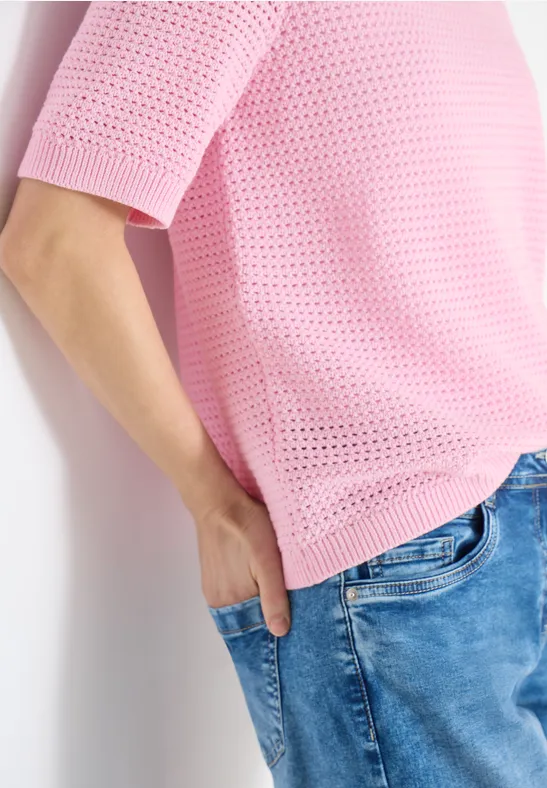 Person im pinken Lochstrick-Pullover mit kurzem Arm, eine Hand in der Hosentasche blauer Jeans