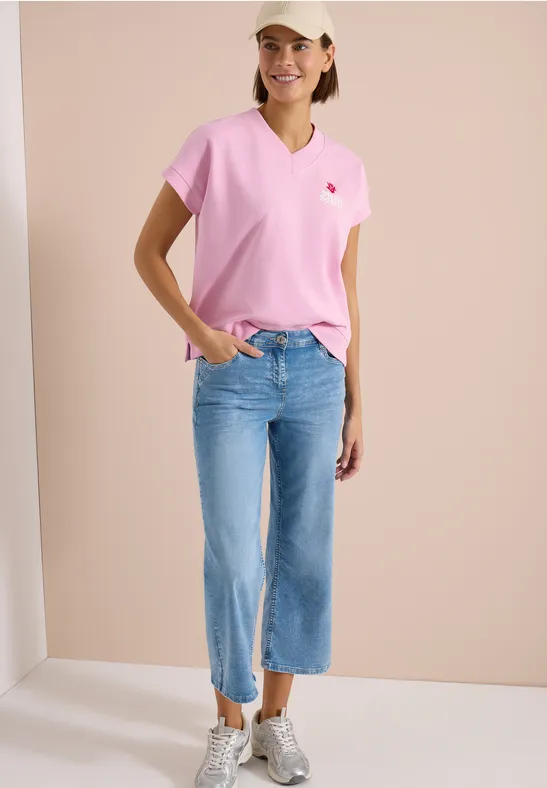 Femme en T-shirt rose, jean bleu clair large coupé et baskets blanches devant un fond beige.