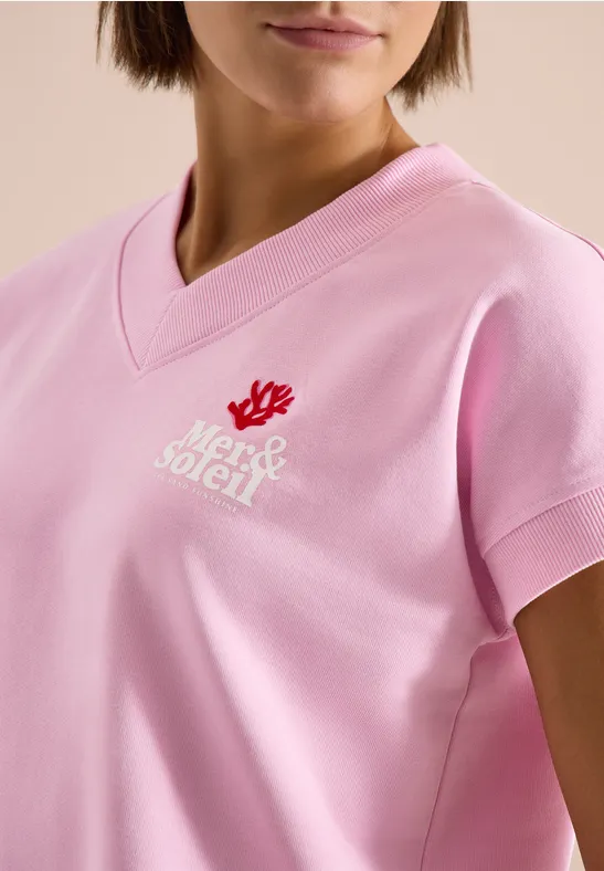Femme portant un t-shirt rose à col V avec un motif de corail rouge et un logo blanc.