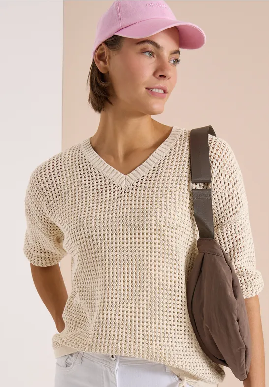 Femme en pull résille, jean blanc, casquette rose et sac épaule sur fond clair.