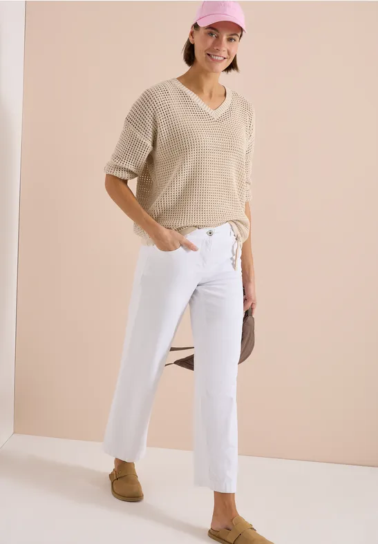 Une personne porte un pull beige à col V, un jean blanc et des mocassins marron.