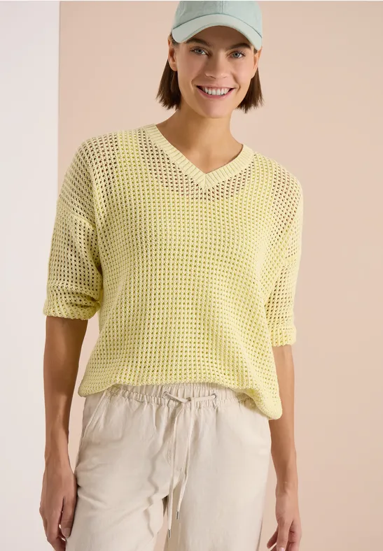 Femme portant un pull maille jaune à col en V et pantalon ample beige sur fond clair.