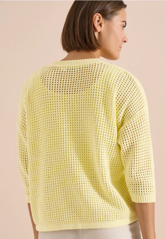 Vue de dos d’une femme portant un pull jaune ajouré ample à manches trois-quarts.