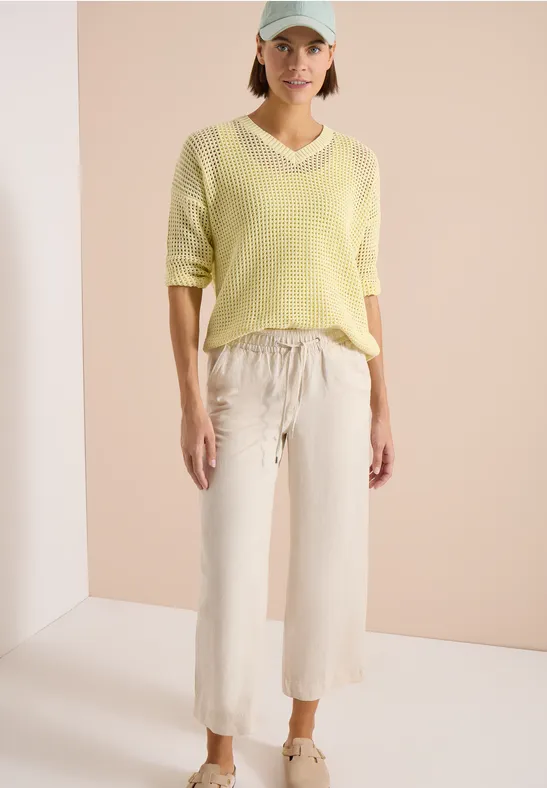 Femme en pull jaune et pantalon clair ample devant un fond sobre.