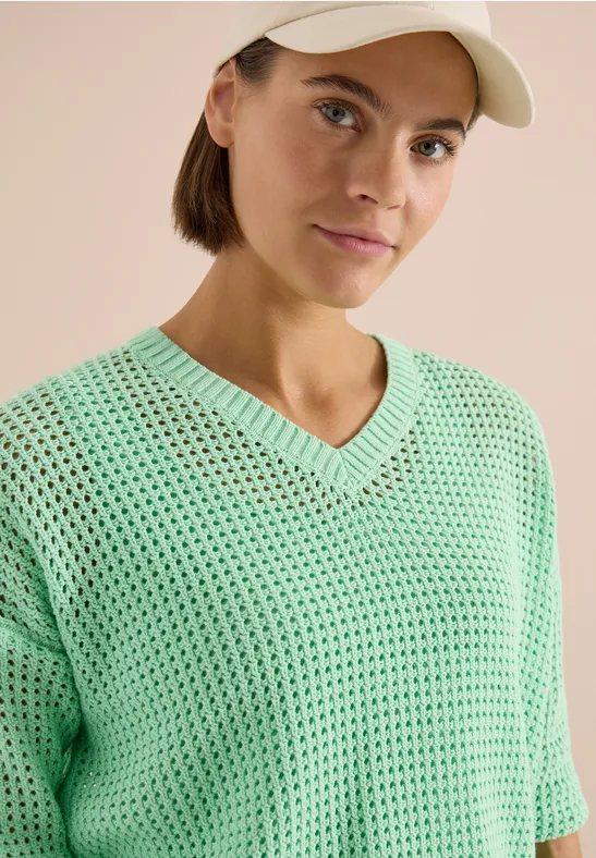 Eine Person mit hellgrünem, locker gestricktem V-Ausschnitt-Sweater und Kappe vor beigem Hintergrund.