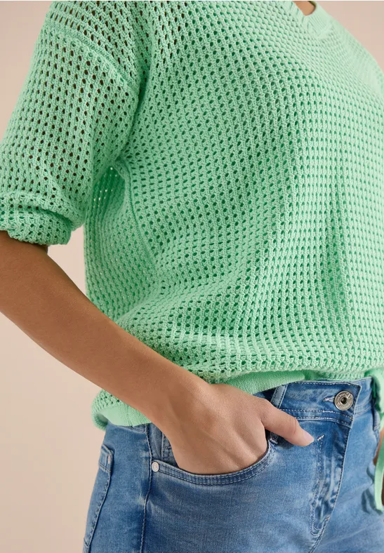 Person mit mintgrünem, locker gestricktem V-Ausschnitt-Pulli, eine Hand in der Jeans-Tasche.