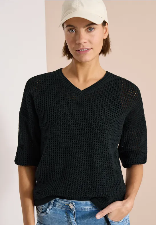 Person in schwarzem, lockerem Strickpullover mit V-Ausschnitt und halblangen Ärmeln, Hand in Jeans-Tasche.