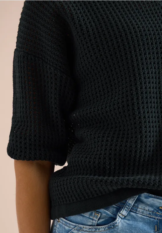 Person in schwarzem grob gestricktem Pullover mit 3/4-Ärmeln und blauen Jeans, nur Oberkörper sichtbar.