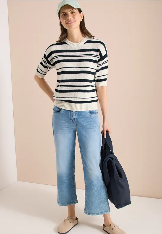 Femme en pull rayé, jean large court et sac sombre sur fond clair.