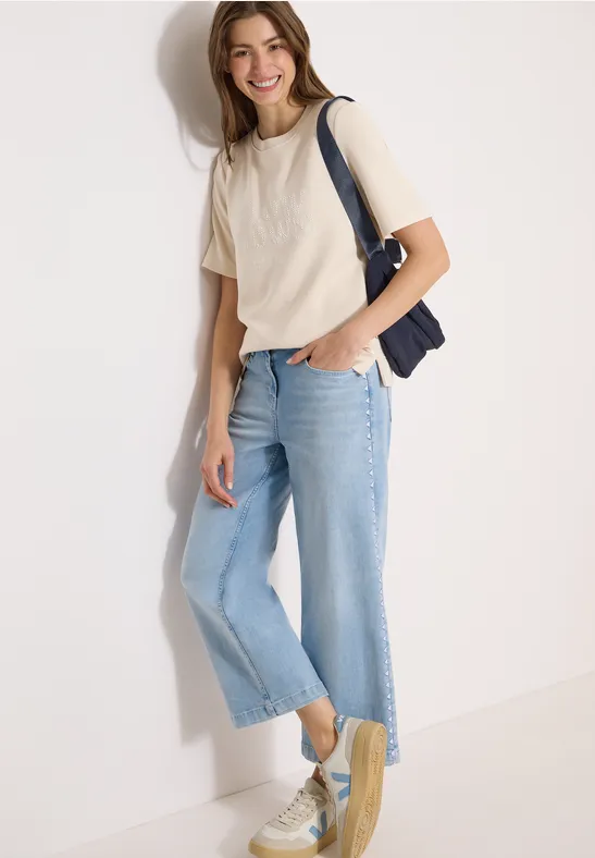 Personne en T-shirt blanc, jean large clair, baskets et sac épaule bleu marine appuyée contre un mur.
