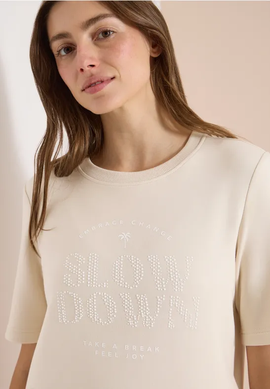 Une femme porte un T-shirt beige clair avec l'inscription « SLOW DOWN » en strass.