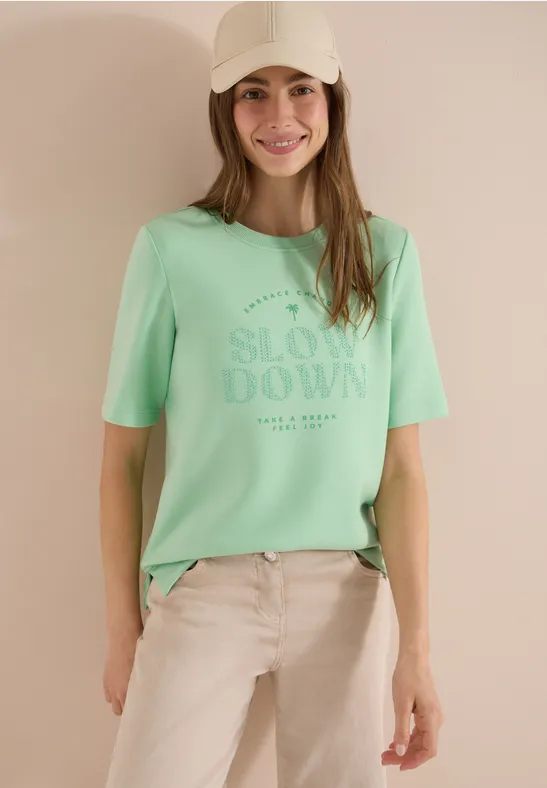 Femme en T-shirt vert pastel avec l'inscription 'SLOW DOWN' devant un fond neutre.
