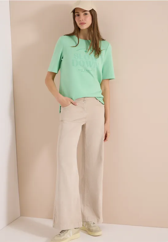 Femme portant un t-shirt menthe et un pantalon large beige sur fond neutre.