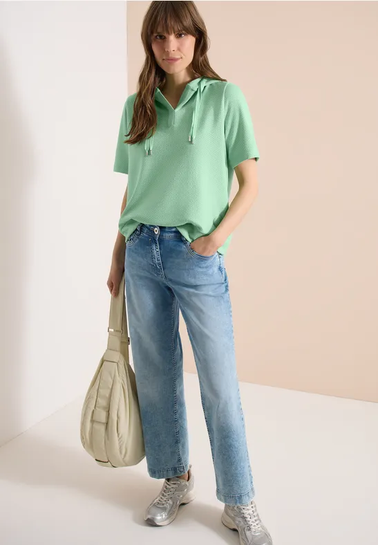 Femme en polo vert, jean droit raccourci et baskets grises, tenant un grand sac souple blanc sur fond clair.