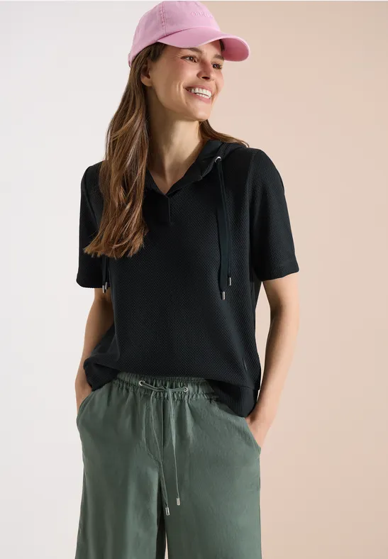 Femme avec casquette rose, sweat à capuche noir à manches courtes et pantalon vert, mains dans les poches.