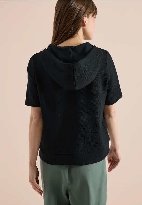 Femme de dos portant un sweat à capuche noir à manches courtes et un pantalon vert sur fond clair.