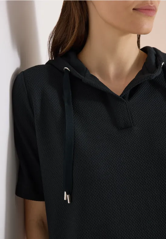 Femme de profil, appuyée contre un mur, portant un t-shirt à capuche noir à manches courtes.