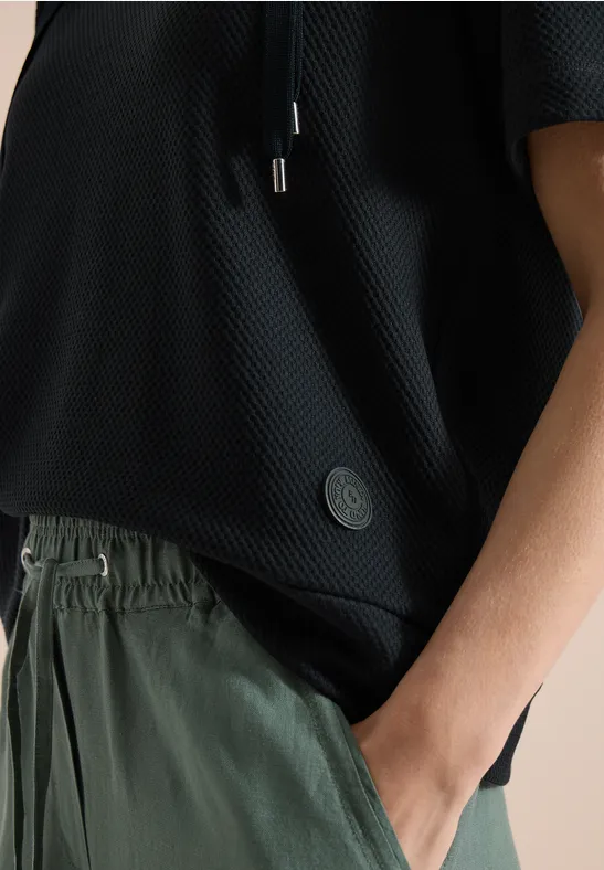Gros plan d'une personne portant un haut noir léger et un pantalon vert, main dans la poche.
