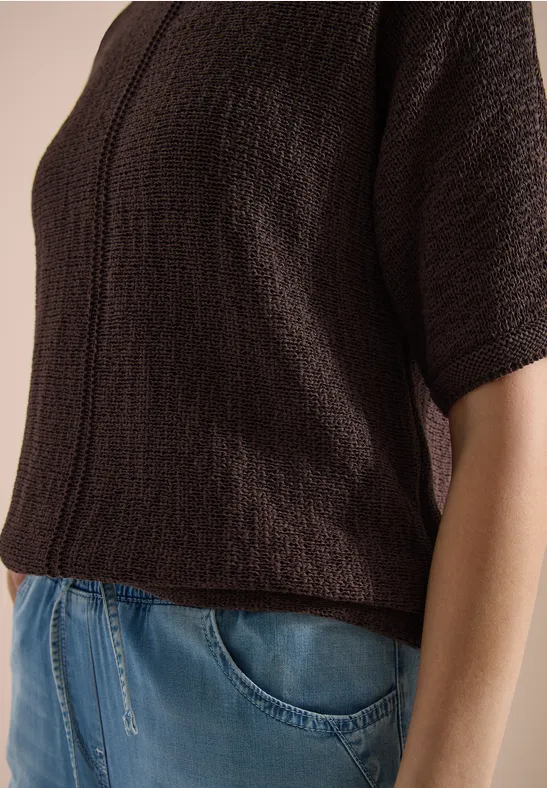 Person trägt braunes, kurzärmeliges Strickshirt zur blauen Jeans.