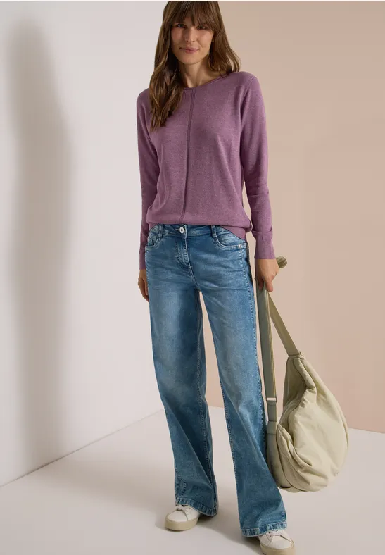 Femme en pull violet, jean bleu et baskets blanches portant un grand sac beige devant un mur clair.