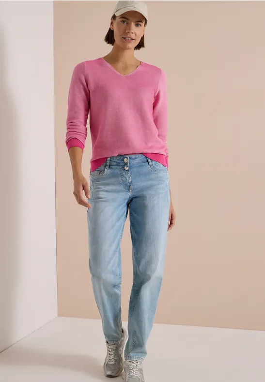 Femme en pull rose à col V, jean clair, casquette blanche et baskets argentées devant un mur neutre.