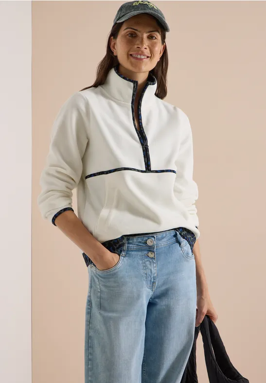 Junge Frau lächelt, trägt weiße Fleecejacke mit Reißverschluss, Jeans und Cap, hält eine Tasche.