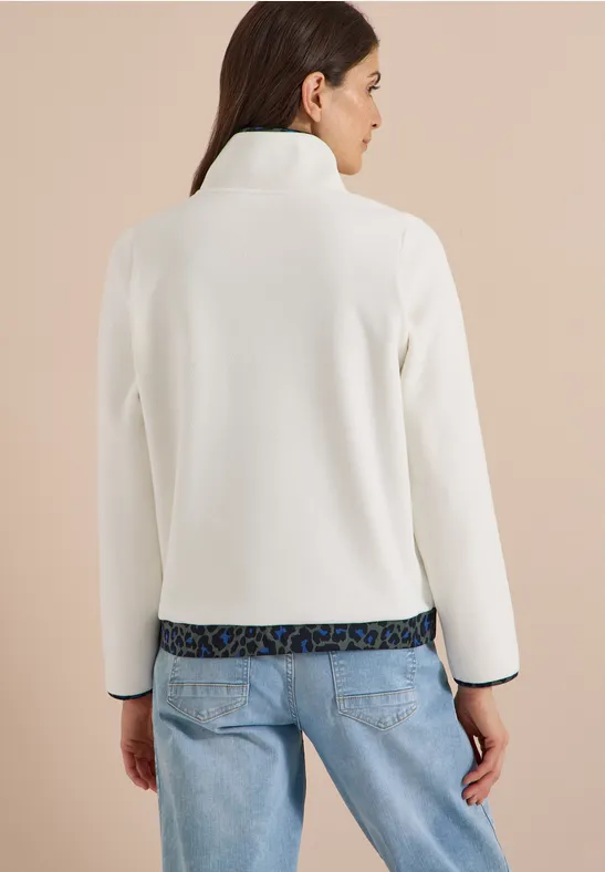 Frau mit Rücken zur Kamera trägt weiße, langärmlige Jacke und blaue Jeans.