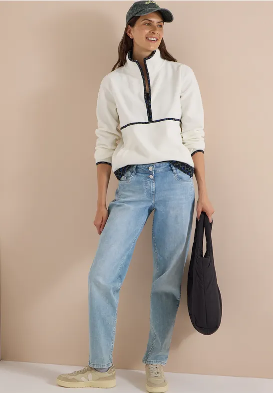 Frau in weißem Fleece-Pullover mit Reißverschluss, hellblauen Jeans, Baseballcap und Tasche.