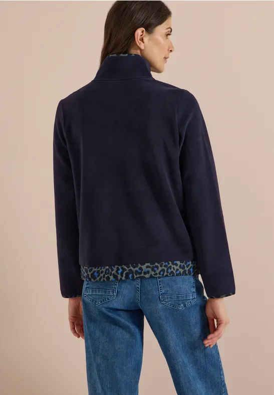 Frau von hinten in dunkelblauer Fleecejacke mit Leoprint-Besatz, Jeans, vor beigem Hintergrund