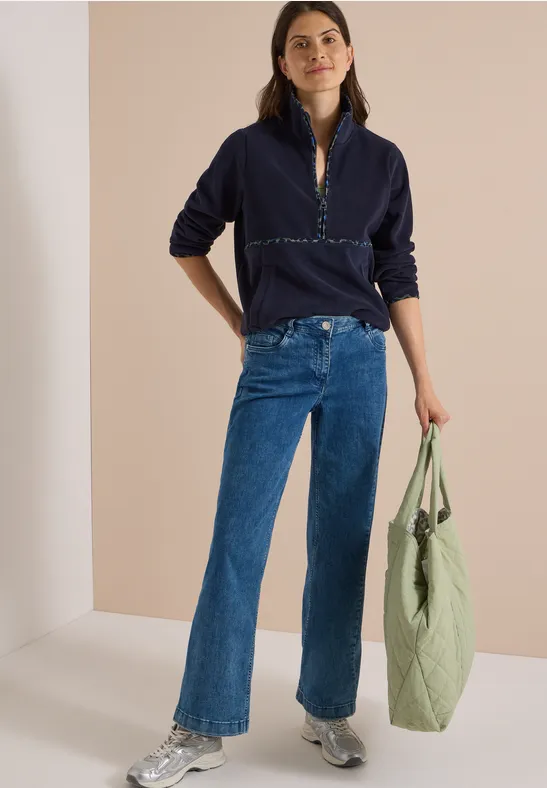 Frau in dunklem Pullover und Jeans trägt eine helle Stofftasche und lächelt entspannt.