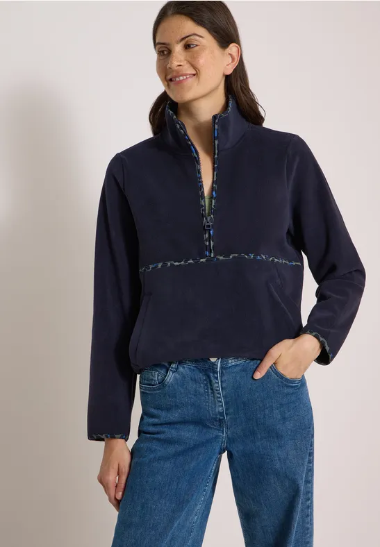 Frau trägt dunkelblaue Fleecejacke mit buntem Zierband an Reißverschluss und Ärmelbündchen, blaue Jeans.