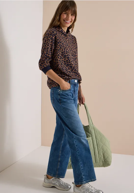 Femme souriante de profil portant un chemisier léopard, jean large, baskets blanches et grand sac vert.