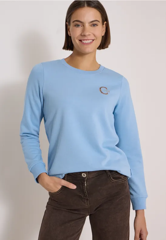 Eine Frau in hellem Langarmshirt und brauner Cordhose posiert vor hellem Hintergrund.