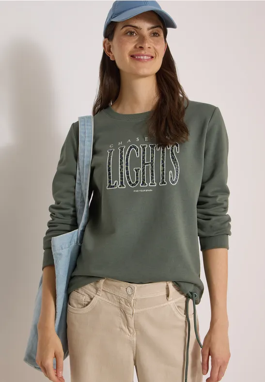 Frau lächelt, trägt ein olivgrünes Sweatshirt mit Aufschrift 'LIGHTS', helle Hose, blaue Cap und Tasche.