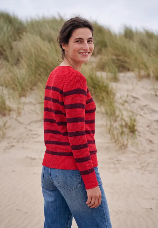 Eine lächelnde Frau in einem rot-schwarz gestreiften Pullover steht am Strand vor Dünen.