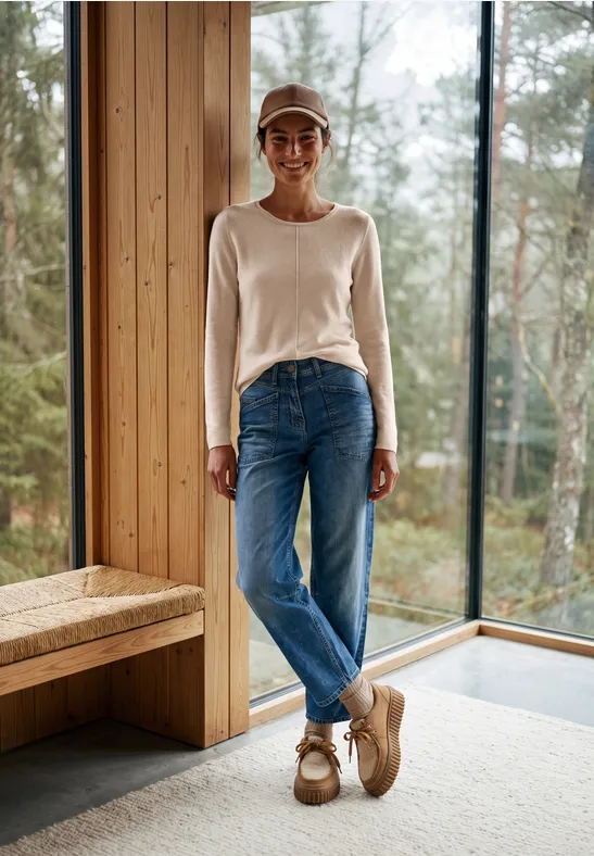 Frau in Jeans und hellem Pullover steht lächelnd in einem Holzhaus vor großen Fenstern, mit Waldblick.