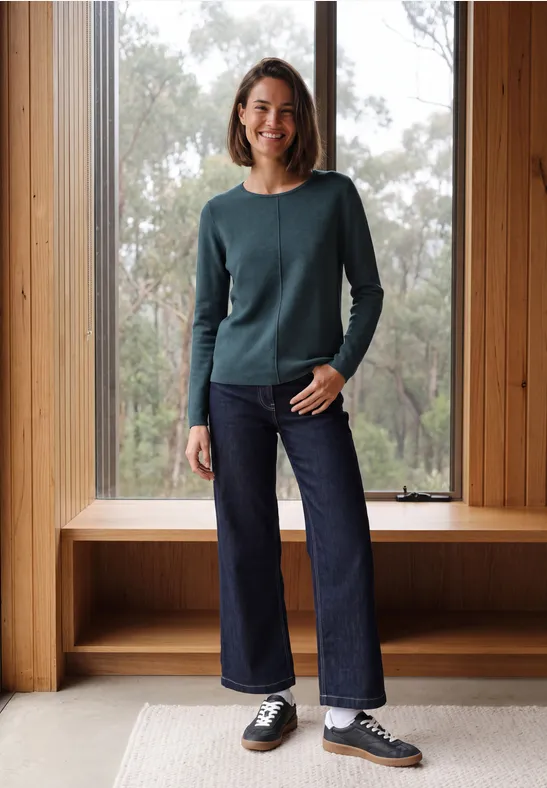 Une femme souriante posant dans un cadre de fenêtre en bois, pull vert, jean sombre et baskets.